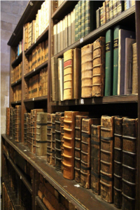 Rows of antique, leather-bound books on dark wooden shelves in a historic library; the books are worn with age and vary in size and thickness.