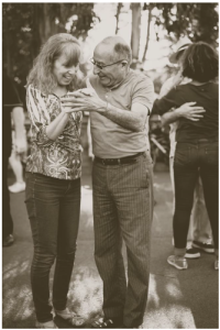 A sepia-toned photograph captures an older man and a woman joyfully dancing together outdoors. The woman, wearing a patterned blouse and jeans, laughs while the man, dressed in a short-sleeved shirt and striped pants, smiles warmly as he guides her in the dance. They are holding hands, with the man leading. In the background, another couple is also dancing, adding to the lively and social atmosphere of the scene. The setting appears to be an outdoor gathering or festival.