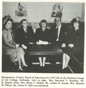 Black and white photo of five members of the Montgomery County Board of Education for 1947–48 seated in the Student Lounge at Montgomery College’s Bethesda campus. The individuals, dressed in formal attire, are seated around a small table beneath two framed prints.