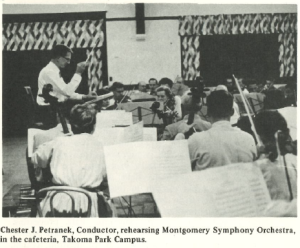 Black-and-white photo of conductor Chester J. Petranek leading a rehearsal of the Montgomery Symphony Orchestra. The musicians, seated with instruments and sheet music, are rehearsing in a cafeteria at the Takoma Park Campus, with patterned curtains visible in the background.