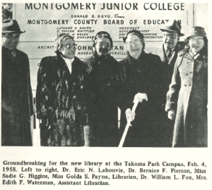 Black-and-white photo of six people standing outside in front of a Montgomery Junior College construction sign during the 1958 library groundbreaking ceremony at the Takoma Park Campus. The group includes college administrators and library staff, bundled in winter coats and smiling, with one person holding a ceremonial shovel.