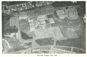 Black-and-white aerial photo of Montgomery College’s Rockville Campus in Fall 1969. The image shows numerous academic buildings, large parking lots filled with cars, athletic fields including a track and tennis courts, and adjacent wooded areas. Roads encircle the campus, with surrounding residential neighborhoods partially visible.