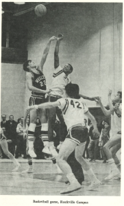 Black-and-white photo of a basketball game at Montgomery College’s Rockville Campus. Two players are mid-air during the tip-off, one from Wesleyan in jersey #22 and the other in a white jersey, while other players and spectators are visible in the background.