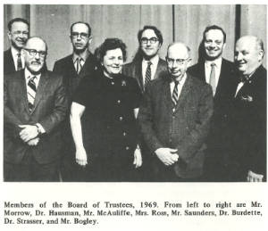 Black-and-white photo of eight members of the 1969 Montgomery College Board of Trustees standing in front of a curtain. Four men stand in the back row, with three men and one woman, Mrs. Ross, in the front. All are dressed in formal attire and smiling.
