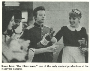 Black-and-white photo of student performers in costume during a scene from Der Fledermaus at Montgomery College’s Rockville Campus. One student in a uniform-style outfit holds a bottle while interacting with others, including a smiling performer in a maid costume.