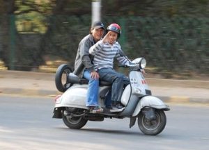Two people ride a white scooter on a road; the driver waves while the passenger sits behind.